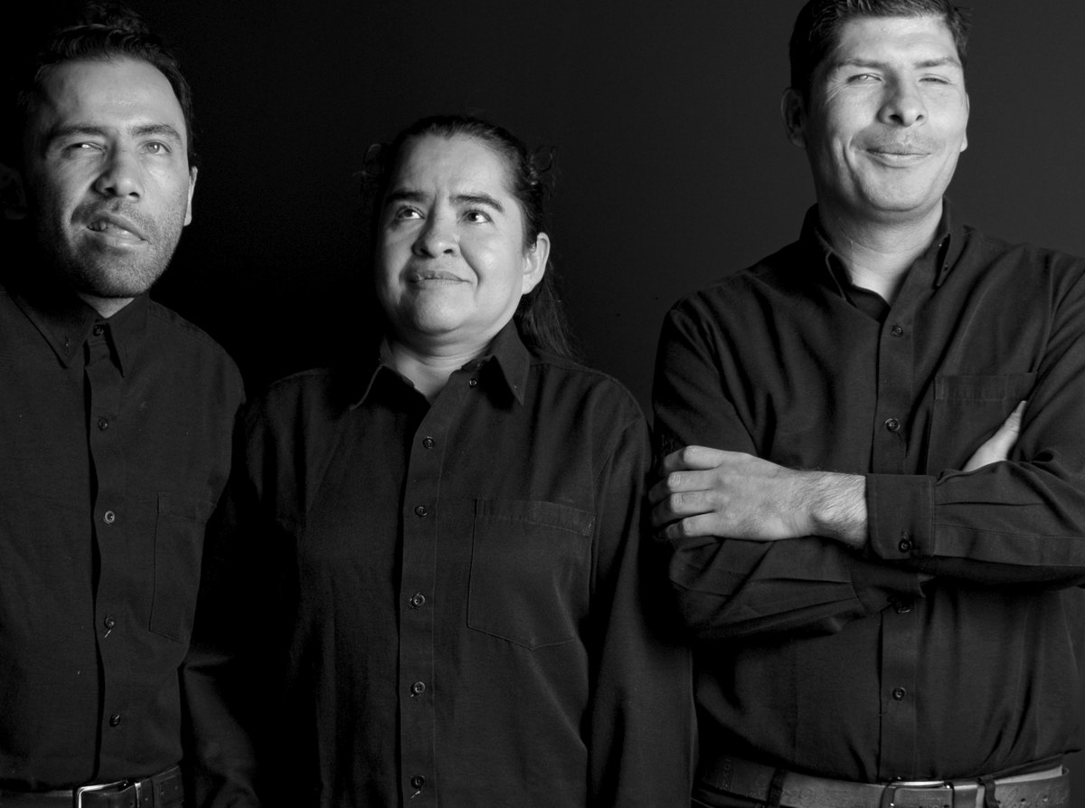 Tres anfitriones de Sombras, vestidos de negro, mirando a distintos puntos de la sala.